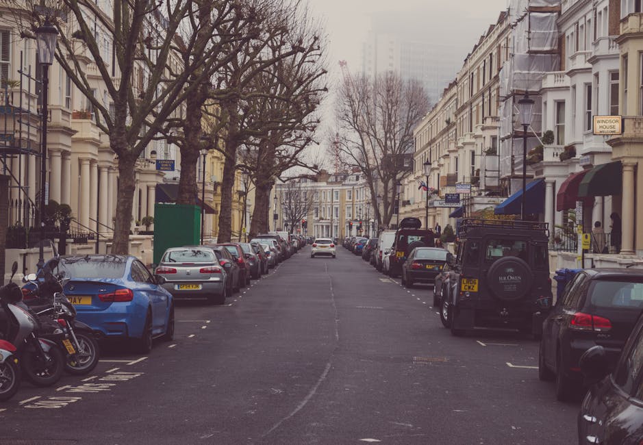 A residential street in Crouch End N8 with closely positioned Victorian-style terraced houses featuring white facades and decorative window trims. Several leafless trees line the pavement, indicating a colder season. Parked cars, including a blue sports car, silver sedans, and a black off-road vehicle, occupy both sides of the street, with clear delineations for parking spaces. Some vehicles are partially behind street furniture such as lampposts and signposts. The road surface is dark asphalt with faint markings, and a white van is visible further down the street. The scene shows a typical urban setting for house removals or furniture transport, with the vehicles either parked or in the process of loading or unloading near the houses. Occasionally, moving equipment such as trolleys or blankets might be used nearby, and the environment is illuminated by overcast natural light, providing a neutral and professional context for house removals in the area, as managed by Man with Van Crouch End.