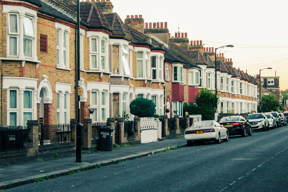 A row of traditional terraced houses with brick facades and bay windows lining a residential street in Crouch End. In the foreground, several cardboard boxes and plastic-wrapped furniture pieces are stacked on the pavement, indicating packing for a home relocation. A person, partially visible, is lifting or arranging items near a large, open cardboard box, preparing for furniture transport. To the right, a white van from Man with Van Crouch End is parked on the street, ready for load-in, with additional vehicles parked behind it. The scene shows the loading process as part of furniture transport and packing and moving activities, with a street lamp and a few trees interspersed along the sidewalk, under warm natural lighting typical of early evening. This setup reflects a professional removals service preparing a house for relocation in Crouch End.