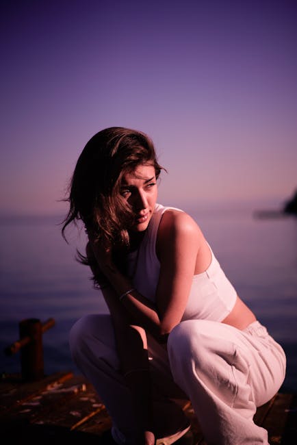 A woman wearing a white sleeveless top and light-colored pants crouches outdoors near a body of water during twilight, with a soft, purple-tinged sky in the background. She is positioned close to a wooden dock or platform, with her left hand resting on her knee and her right arm resting on her thigh. Her dark hair falls loosely around her face, and she gazes downward with a contemplative expression. In the background, blurred elements of the water and distant shoreline are visible, with subtle natural lighting giving a calm, serene atmosphere. The scene suggests a moment of pause during an outdoor setting, with no visible furniture or moving equipment present.