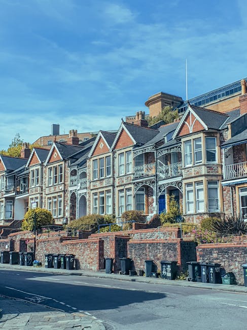 A row of traditional terraced houses with brick facades and bay windows lining a residential street in Crouch End. In the foreground, several cardboard boxes and plastic-wrapped furniture pieces are stacked on the pavement, indicating packing for a home relocation. A person, partially visible, is lifting or arranging items near a large, open cardboard box, preparing for furniture transport. To the right, a white van from Man with Van Crouch End is parked on the street, ready for load-in, with additional vehicles parked behind it. The scene shows the loading process as part of furniture transport and packing and moving activities, with a street lamp and a few trees interspersed along the sidewalk, under warm natural lighting typical of early evening. This setup reflects a professional removals service preparing a house for relocation in Crouch End.