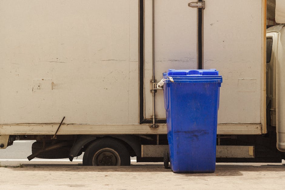 The image shows the side view of a white delivery van with a closed rear door, parked on a paved street. A large blue recycling bin with a closed lid is positioned next to the van's rear, secured with white straps, indicating it is ready for collection or loading. The bin appears to be made of plastic and is placed on the pavement directly beside the van, which has a small visible wheel and part of the undercarriage. The scene likely captures a moment during a home relocation or waste collection process, with the van possibly used for furniture transport or packing and moving tasks in a house removal service. The background is plain, focusing on the loading area, and the lighting suggests daytime, with natural light illuminating the objects clearly. The image emphasizes the logistics of moving and waste disposal associated with house removals in the Crouch End area, relevant to the services provided by Man with Van Crouch End.