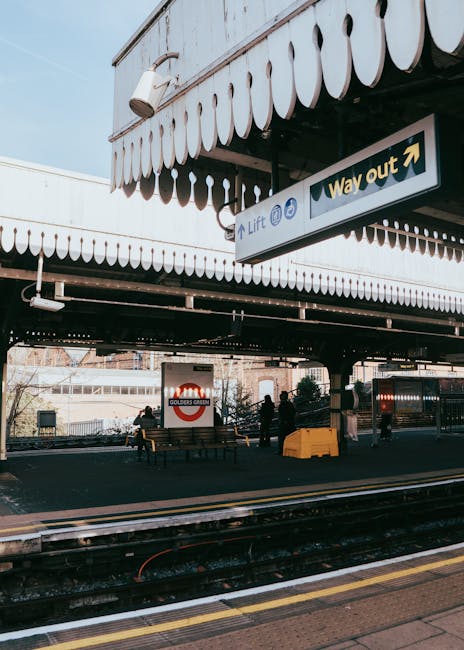 The image shows a train station platform with a concrete overpass above, featuring a white scalloped edge canopy and a 'Way out' sign indicating the exit. Beneath the overpass, there are several peopl