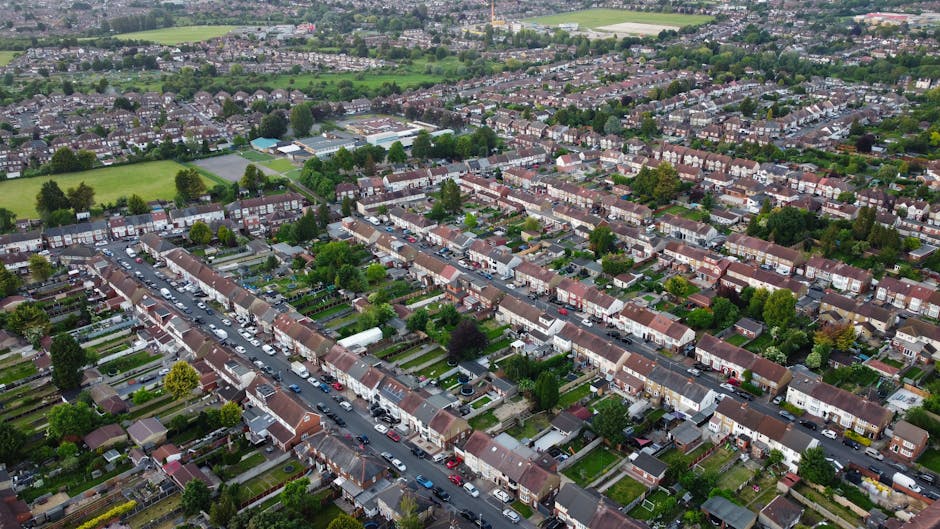 Aerial view of a residential neighbourhood during daytime showing a row of terraced houses with small front gardens, parked cars lining the streets, and houses with pitched roofs and brick facades. Some properties have driveways or backyards visible, with a few home furniture or moving boxes seen near the entrances. The area is lush with numerous trees and greenery. In the background, there are open green fields and recreational areas. The image captures a typical suburban street scene that might be relevant to house removals or home relocation services, illustrating the context of vehicle loading and moving preparations undertaken by a company such as Man with Van Crouch End for local moves.