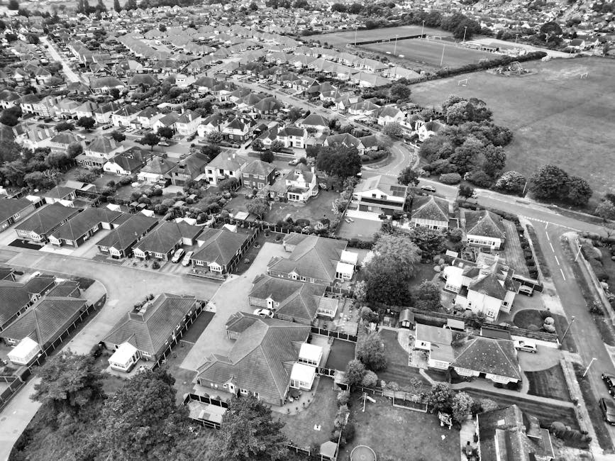 An aerial black-and-white photograph of a residential neighbourhood showing multiple houses with pitched roofs, some with small gardens or yards at the front and back. Several properties are connected by driveways and pathways, with parked cars visible along the streets and in driveways. The image captures a variety of building materials, including brick and tile roofing, and some houses have visible porches or small extensions. Trees and shrubs are interspersed throughout the area, providing greenery amid the densely packed homes. In the background, open sports fields and larger green spaces are seen adjacent to the residential area, with a main road visible at the edge of the neighbourhood. The overall scene suggests a quiet, suburban environment during daytime, with no active moving process visible in the image. The photo, associated with house removals and moving services, highlights the typical layout of homes that a company like Man with Van Crouch End might service within the area, supporting home relocation and furniture transport activities. 