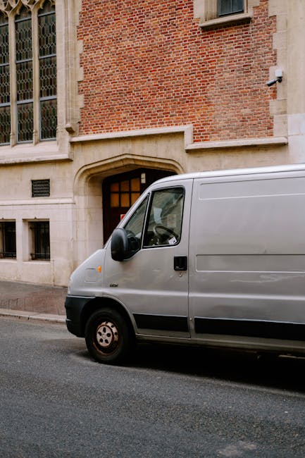 A grey moving van parked directly in front of a historic brick building with large, multi-pane stained glass windows and an arched stone doorway. The van's side door is closed, and it is positioned on a narrow street with a paved surface. The building's exterior features a combination of red brick and light stone detailing around the windows and entrance. The scene captures part of the van's front and side, with a focus on the loading area near the building's entrance, indicating a home relocation or furniture transport process in progress, typical of local house removals services offered by Man with Van Crouch End.