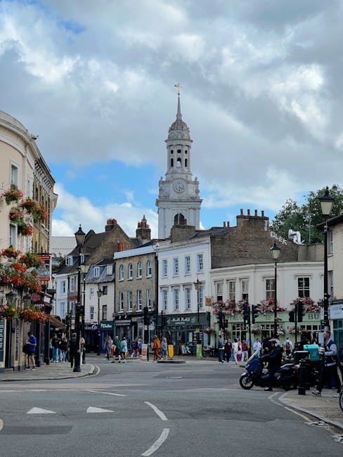 A busy street scene in Crouch End with pedestrians walking along the pavement, some carrying shopping bags. On the left, there are multiple shops with large windows, including a florist with hanging baskets of flowers and a clothing store. Several vehicles, including motorcycles and cars, are parked along the roadside, and a black van is visible in motion. The street is lined with black lampposts decorated with flower boxes. In the background, historic buildings with classic architecture, including a prominent white clock tower with a spire topped by a golden weather vane, rise against a partly cloudy sky with patches of blue. The scene captures an active, urban environment typical of a residential and commercial district in Crouch End, suitable for house removals and moving services with clear indications of loading and transport logistics taking place nearby.
