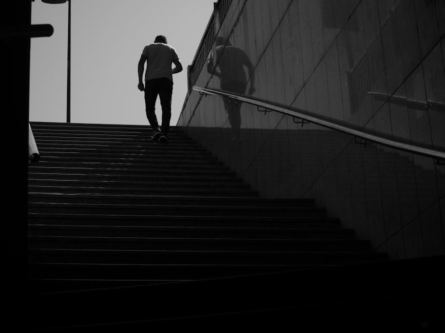 A black-and-white photograph showing a person wearing a short-sleeved shirt and dark trousers ascending a set of outdoor stairs with evenly spaced steps, leading to an upper level of a modern building. The stairs are made of a dark material, possibly metal or wood. The individual is carrying nothing in their hands, and their reflection is visible on the sleek, glass-panelled wall on the right side of the image, which is supported by a metal railing running parallel to the steps. The surrounding environment appears urban, with a minimalistic architectural style, and the sky above is overcast, providing diffuse lighting. This scene illustrates the physical effort involved in stairway navigation during home relocations or furniture transport steps, where careful planning is required to manage movement restrictions like staircase restrictions on Tottenham Lane. Man with Van Crouch End occasionally services such property access challenges during removals or moving logistics, including stairway handling and loading procedures.