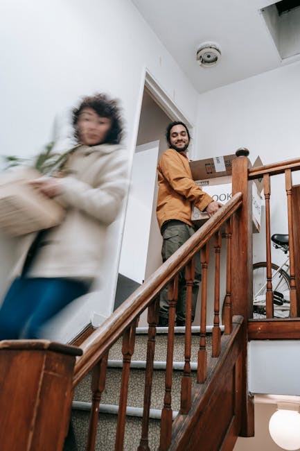 Inside a residential property during a house removal, a man with a beard, wearing a brown shirt and grey trousers, is standing at the top of a staircase, smiling while carrying a box labeled 'kitchen.' A woman, dressed in a white jacket and blue jeans, is walking past the stairs with a blurred motion indicating she is moving quickly, possibly carrying a small item. The staircase has wooden banisters and carpeting on the steps, and several cardboard boxes, some sealed with packing tape, are positioned nearby, suggesting a packing and moving process. The environment is well-lit with natural light, and visible items such as a bicycle in the background imply a recent or ongoing home relocation. This visual captures part of the loading process typical of furniture transport and packing services offered by Man with Van Crouch End, relating to staircase restrictions on Tottenham Lane and general removals tasks.